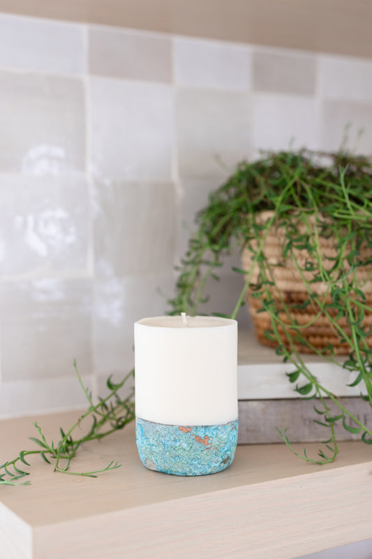 A Coconut Sandalwood pillar candle with a white upper part and a textured blue lower part, placed on a surface with a plant in the background.