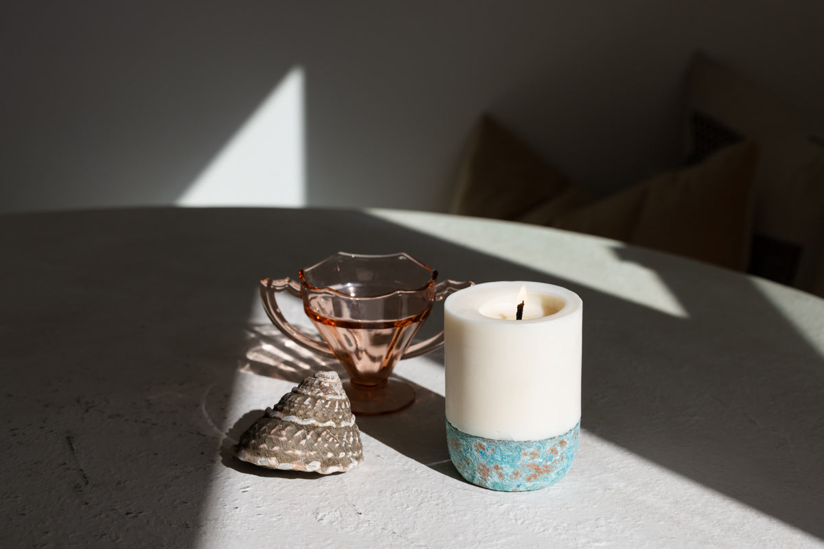 A white artisan pillar candle with a blue patterned base, placed on a weathered-concrete surface, accompanied by a bronze-colored metal cup and a decorative shell, with dappled light from a window. the scent is eucalyptus, bergamot, lemon, sage, fig, rose, leaves