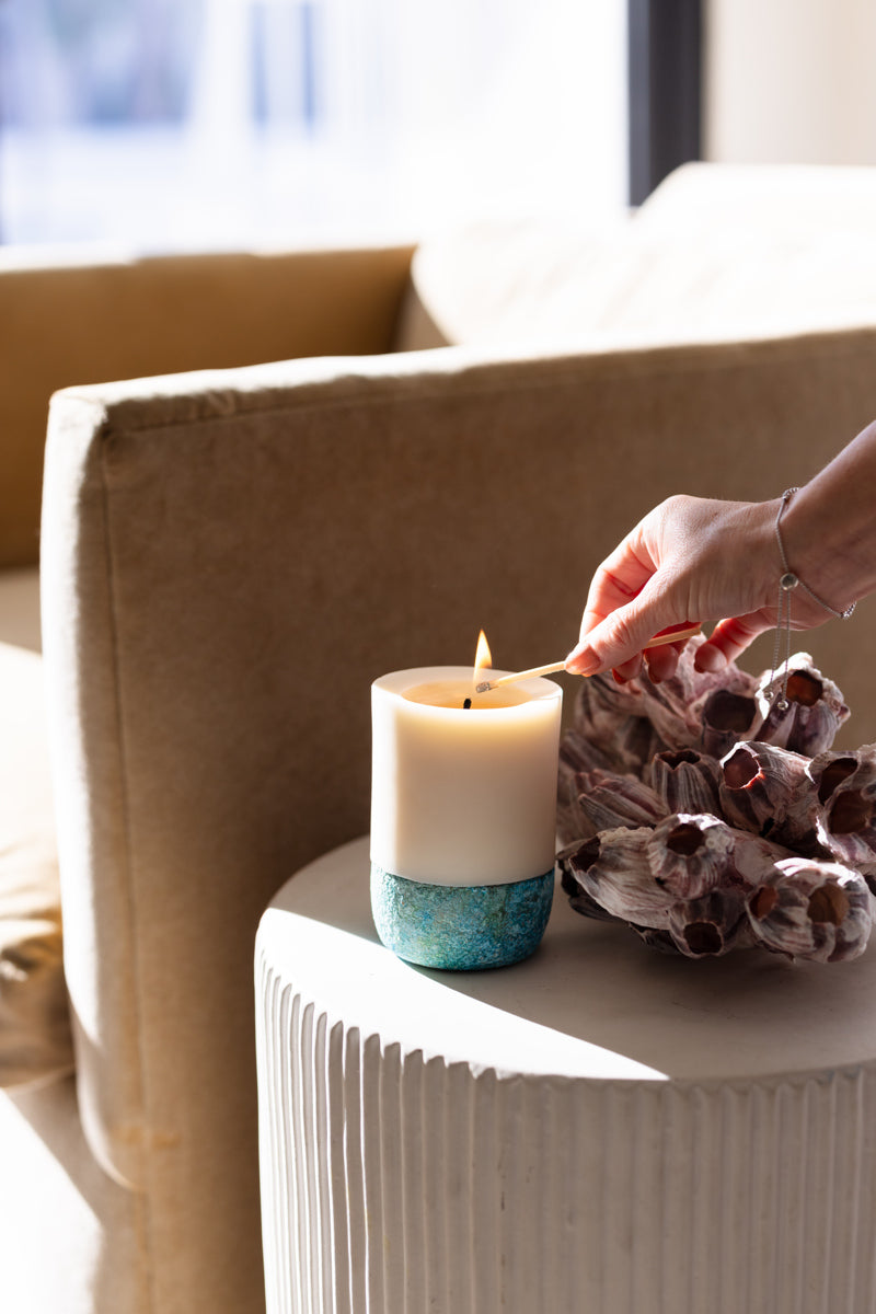 A lit pillar candle with a white wax and a blue and beige design on the base, placed on a white surface, with a person's hand holding a floral arrangement in the background.