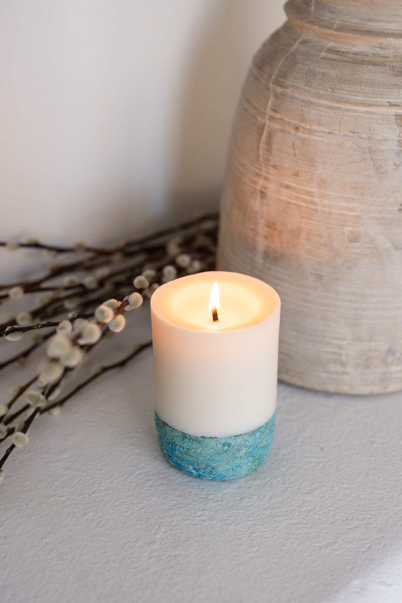 A lit pillar candle with a white upper half and a blue patina-finished concrete base, placed on a gray surface next to a decorative vase and some plant branches.