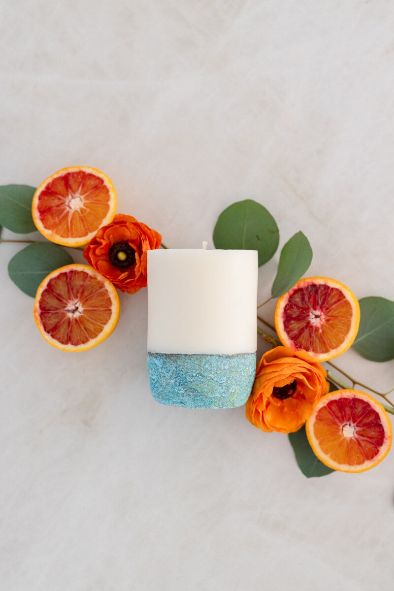 A white pillar candle with a blue and gray mottled base, surrounded by dried orange slices and green leaves on a light background.